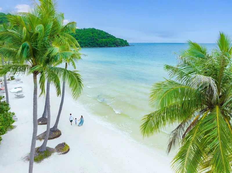 A family walks along a white sand beach fringed by palm trees with turquoise water in Phu Quoc. A family walks along a white sand beach fringed by palm trees with turquoise water in Phu Quoc.